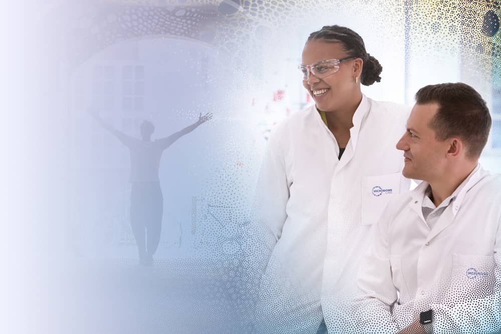 A woman wearing a Microbiome Labs lab coat standing next to a man who is sitting own also wearing a Microbiome Labs lab coat