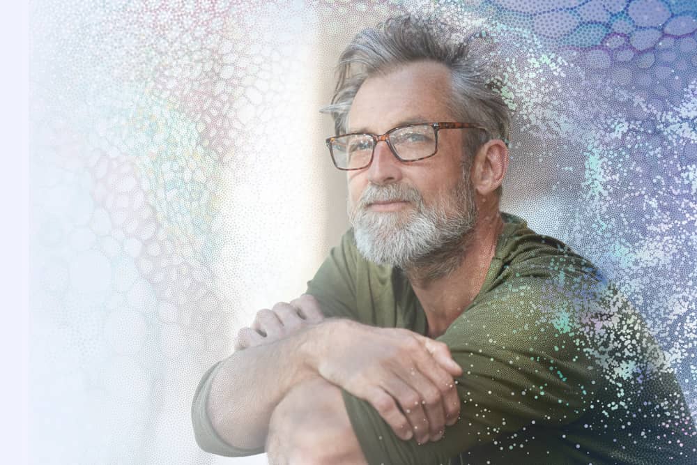 A man with grey hair and a beard wearing glasses is sitting down with his arms folded looking out